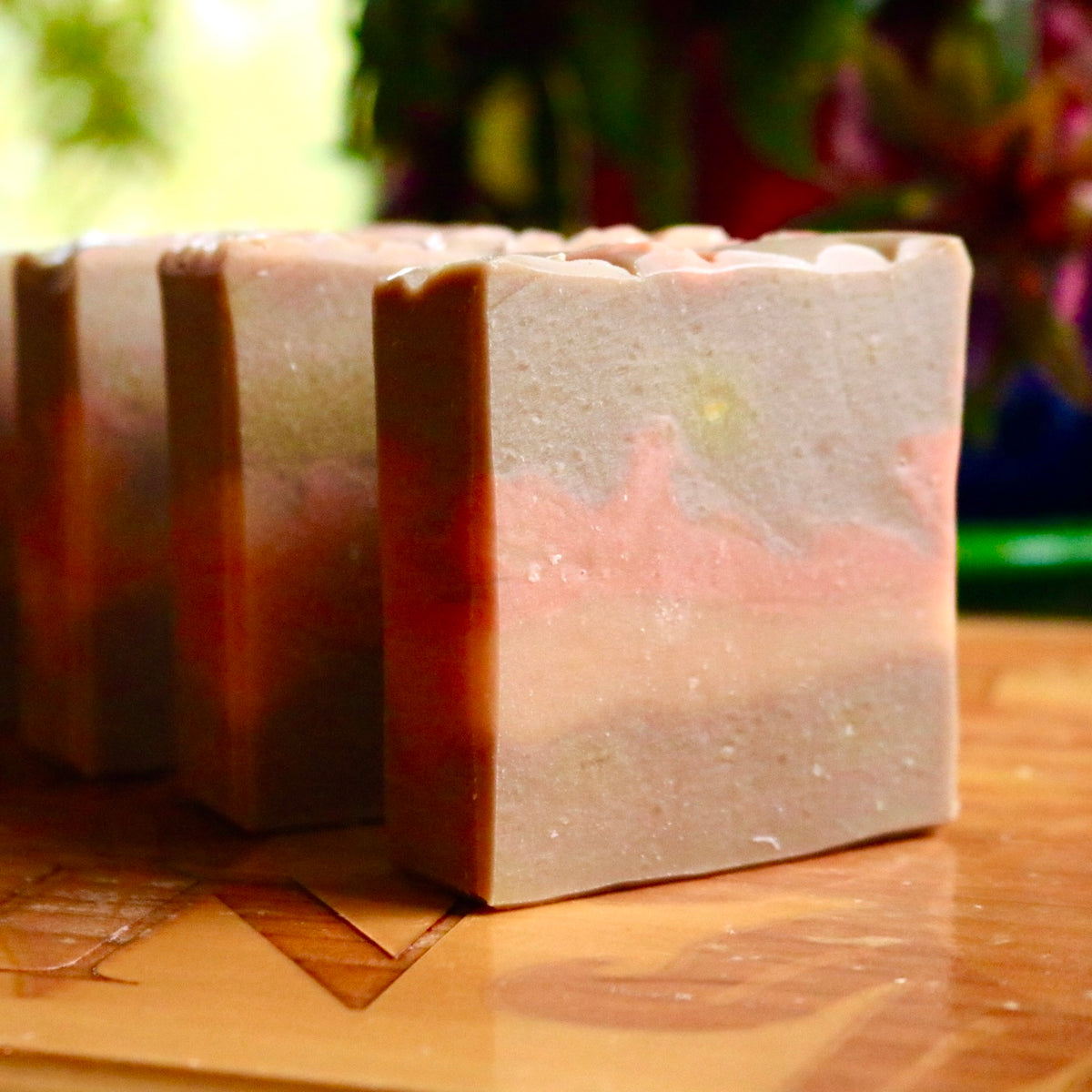Four plant-based Sweet Fig bar soaps seen in a group, with one in front of the other in front of vibrant background of plants and pottery. These bars show different striations of color produced by hand in the cold process method.