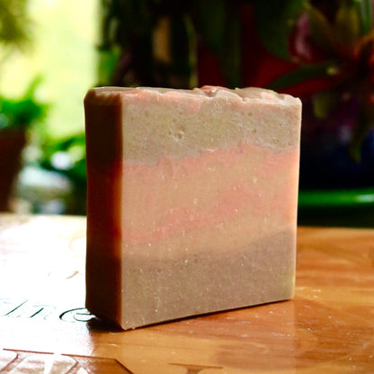 Single bar of Sweet Fig vegan soap, seen atop a wooden base in front of a background of plants in an indoor setting. The bar has a brown top and bottom with pink streaks on an orange middle section.