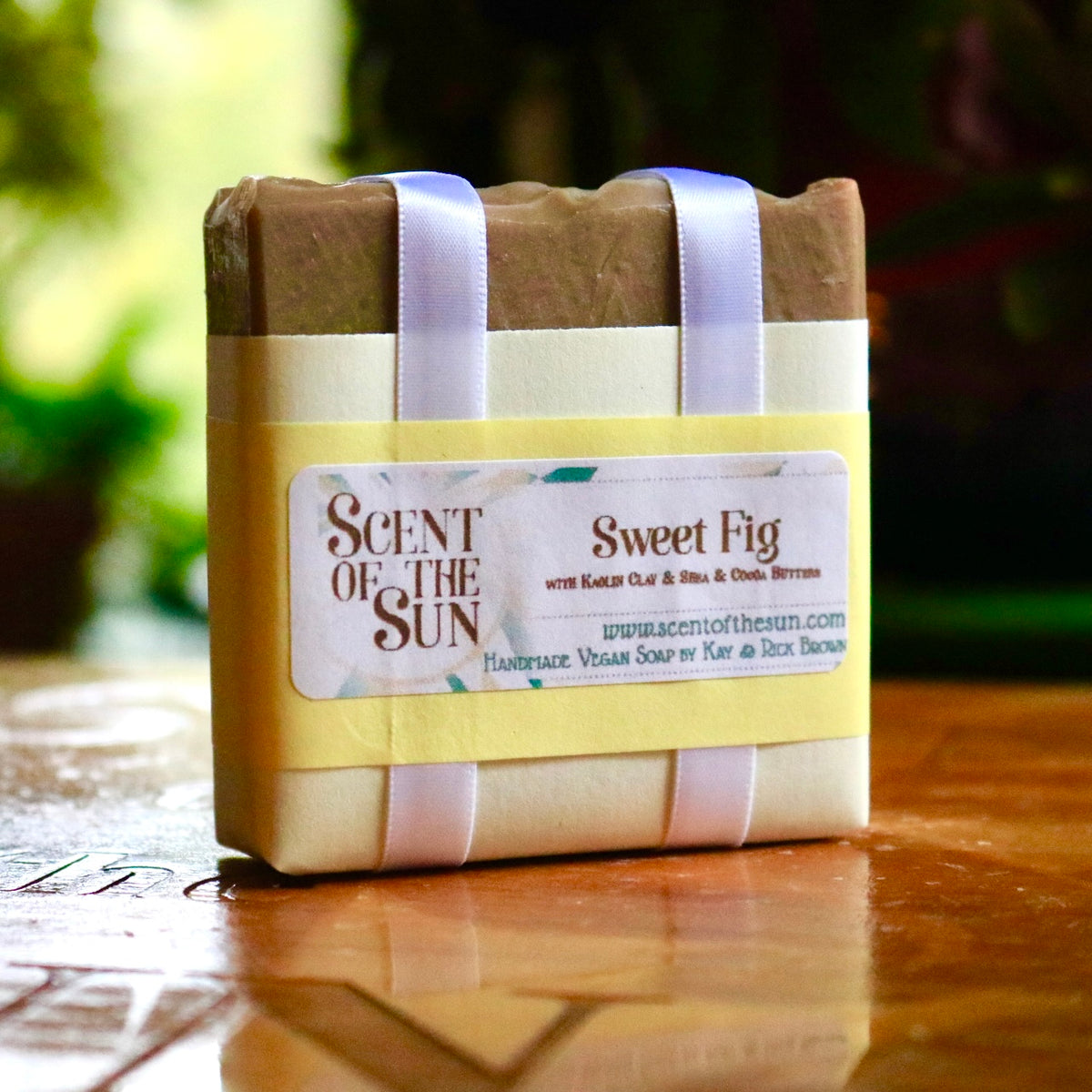 Wrapped bar of Sweet Fig bar soap, a plant-based creation wrapped in white satin ribbon holding together a simple paper covering making the soap appear to be giftwrapped. Text on the label states the soap features kaolin clay, shea butter, and cocoa butter.