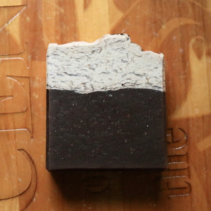 Single bar of vegan Caffe Mocha soap seen laying atop a wooden background. This plant-based soap has a dark brown base and a beige top and is sprinkled at the very top with ground coffee.